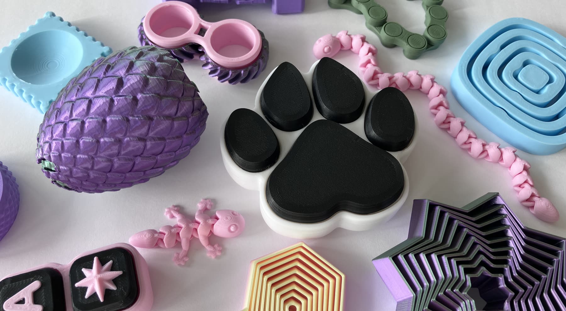 A close spread of Fidget Street fidgets — a black paw clicker, a purple dragon egg, a pink chain, nested shapeshifters and textured cubes
